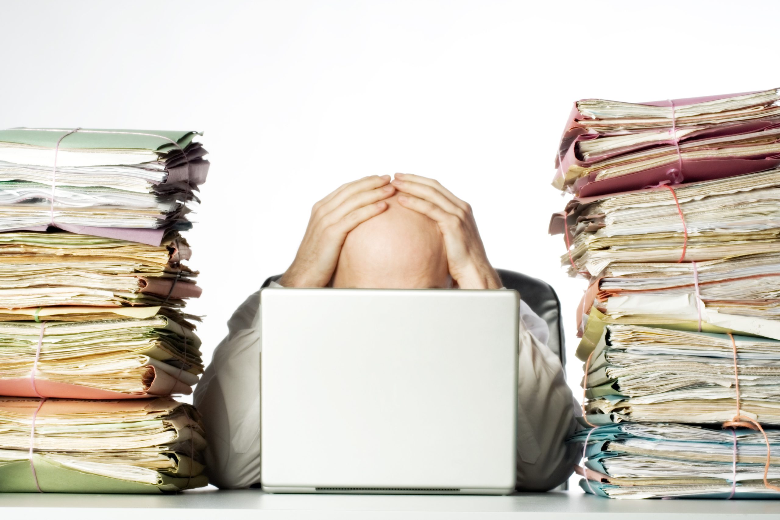 Male businessman sitting behind a laptop, his face hidden, with his hands on top of his head. Two large piles of paperwork are piled on each side of the model, towering over his head. Isolated on white background.